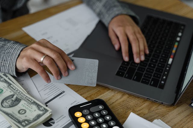 Person using a laptop while holding a bank card with money and a calculator beside them